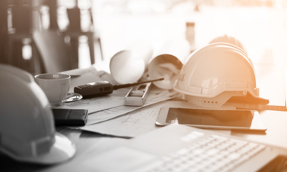 Blog - Hard Hats Sitting on a Desk with Documents, a Keyboard, Coffee Cup, and Other Supplies